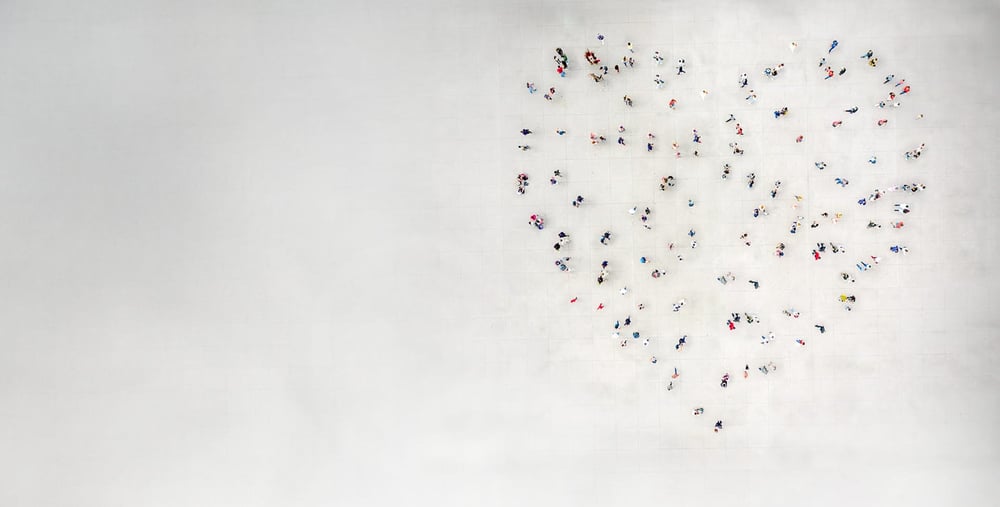 Människor står utspridda så att de tillsammans bildar formen av ett hjärta på en stor öppen yta, sedd uppifrån.