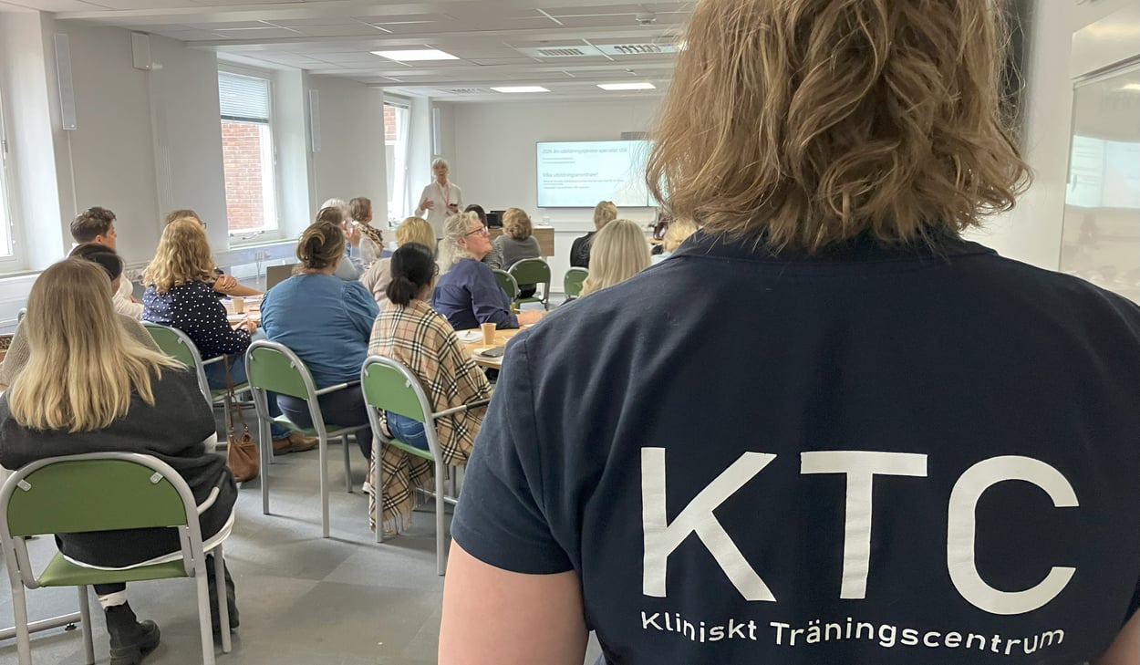 Person med tröja märkt ”KTC Kliniskt Träningscentrum” står i ett konferensrum där deltagare lyssnar på en presentation.