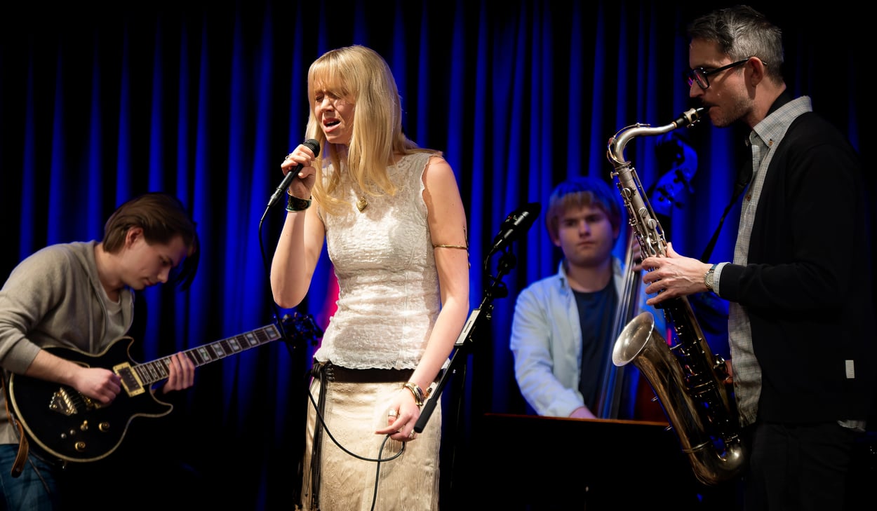 Musiker uppträder på scen med sång, gitarr, kontrabas och saxofon.