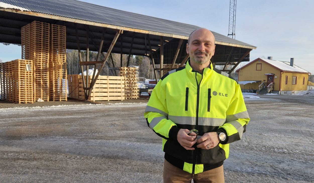 Mattias Ringström iklädd gul varseljacka står på en industriell gårdsplan med staplade träpallar och ett skjul i bakgrunden.