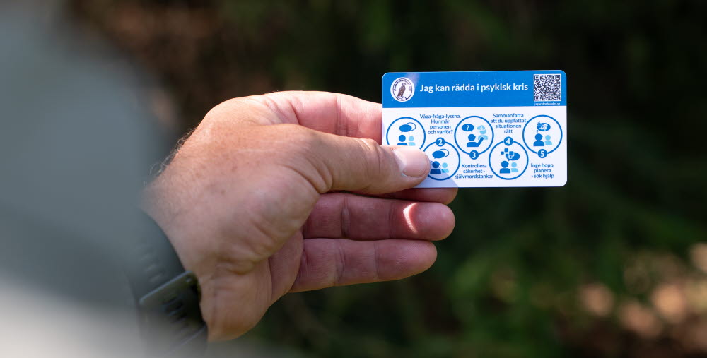 En hand håller ett kort med rubriken "Jag kan rädda i psykisk kris" och fem steg för att hjälpa någon i kris, inklusive att våga fråga, kontrollera säkerhet och söka hjälp.