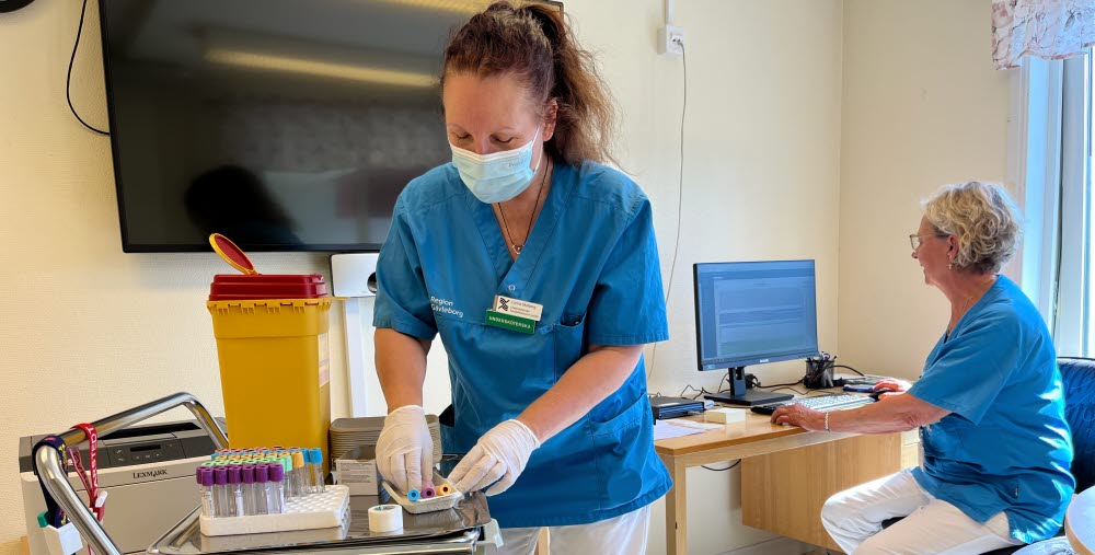 två personer i arbetskläder vid ett rullbord med provrör och vid en dator