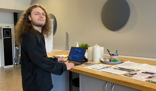 Person vid en dator ståendes vid en arbetsbänk med papper liggandes på sidan.