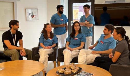 Samlingslokal med vita väggar, två bruna bord, sju personer som sitter och samtalar. Fyra personer i blåa och vita kläder och två personer i privata kläder. AT/BT centrum