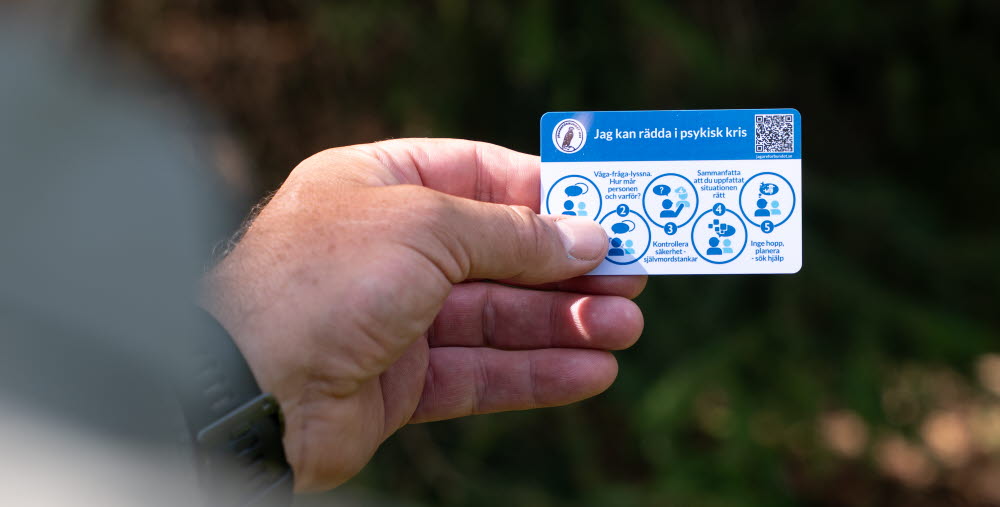 En hand håller ett kort med rubriken "Jag kan rädda i psykisk kris" och fem steg för att hjälpa någon i kris, inklusive att våga fråga, kontrollera säkerhet och söka hjälp.