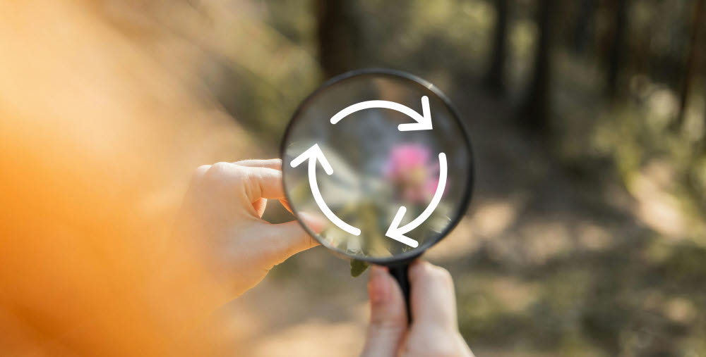 Hand som håller i ett förstoringsglas med skog i bakgrunden. I förstoringsglaset syns en illustration av cirkulär ekonomi