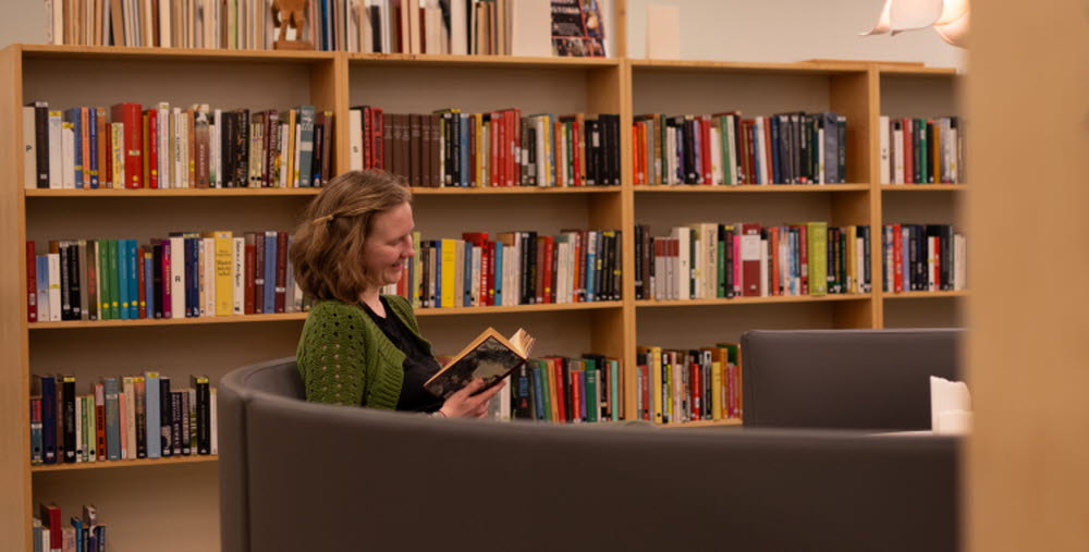 En kvinna i grön tröja sitter i en grå fåtölj i ett bibliotek och läser en bok, med bokhyllor fyllda av böcker i bakgrunden.