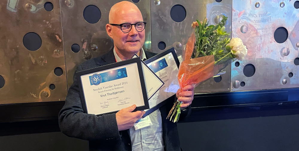 Kärlkirurgen Knut Thorbjørnsen, medelålders man står med blommor i handen och flera inramade prisdokument i handen. Han ler mot kameran. Han bär en ljus skjorta och en grå kavaj. 